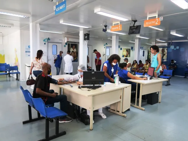Na recepção, o ar-condicionado em funcionamento, luzes acesas, médicos atendendo, pacientes e acompanhantes identificados, bebedouro e ouvidoria